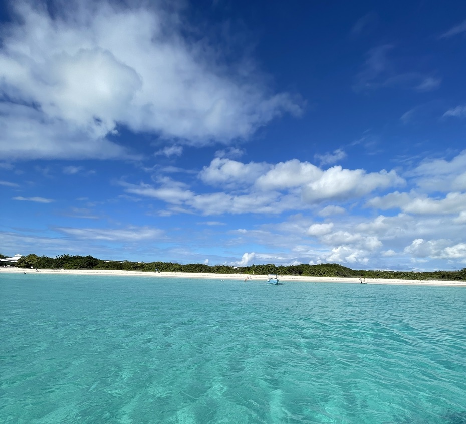 Clear turquoise water stretching toward the long white-sand shore of Rendezvous Bay, Anguilla, under a bright blue sky with drifting clouds.