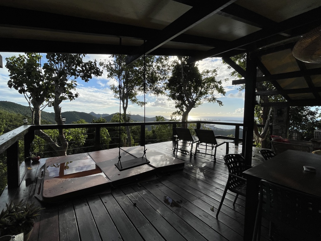 A covered hillside deck with a hanging wooden swing, lounge chairs and a dining table, looking out through trees to green hills and the sea beyond St. Martin.