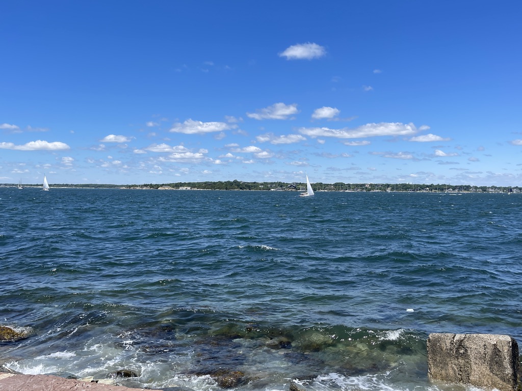 Sailboats on Narragansett Bay under a bright summer sky, seen from the rocky shoreline at Fort Adams State Park.