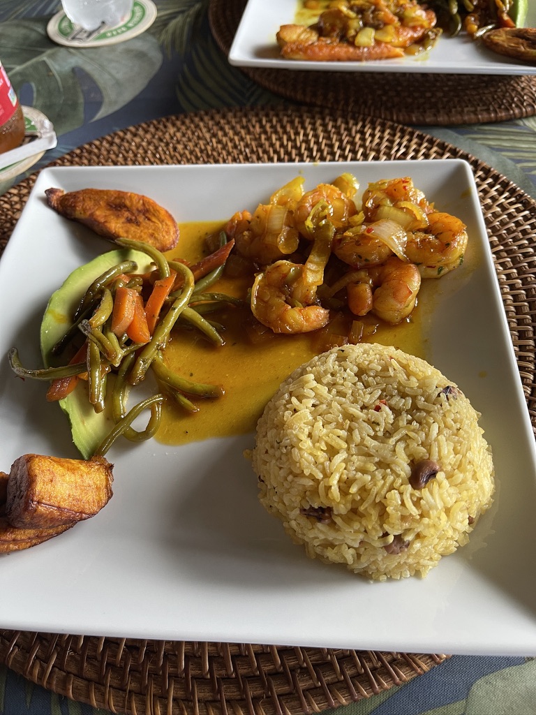 Shrimp in a golden curry sauce with rice and peas, fried plantains, avocado and green beans, served on a white plate over a woven placemat.
