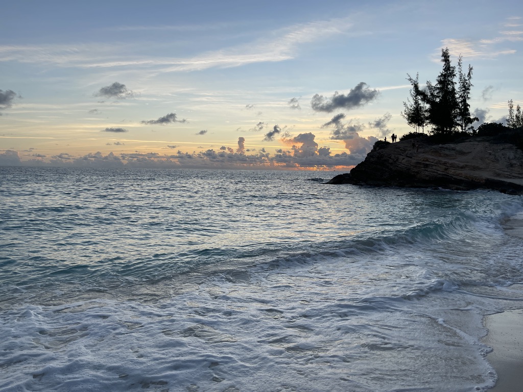 Waves washing onto the sand at Mullet Bay at sunset, with a rocky point topped by pines silhouetted against a soft orange and blue sky.