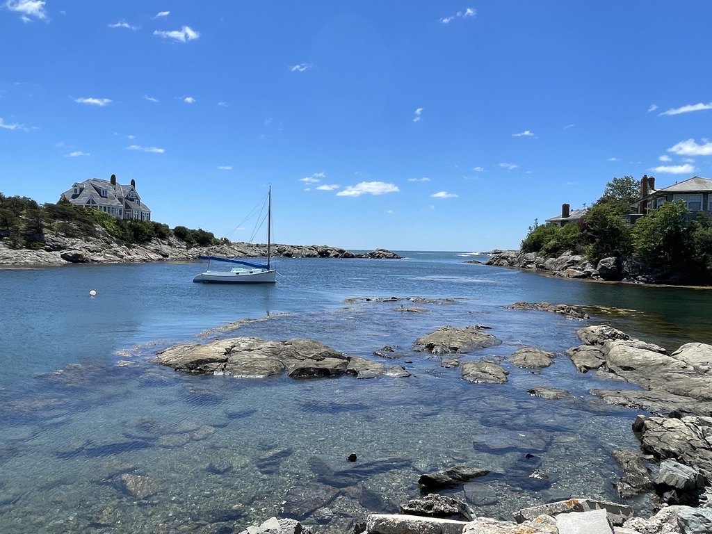 A sailboat moored in a clear, rocky cove along the Newport coastline under a bright blue sky.
