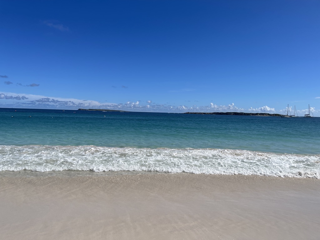 Pale sand and a line of white surf meeting turquoise water under a deep blue sky, with low islands and sailboats on the horizon at Orient Bay, St. Martin.