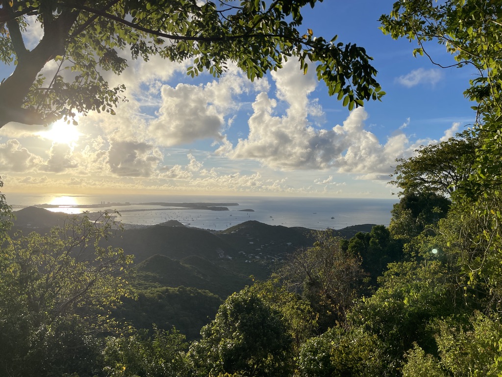 Late sun breaking through the trees above St. Martin's green hills, with the bay, anchored boats and the coastline stretching out below.
