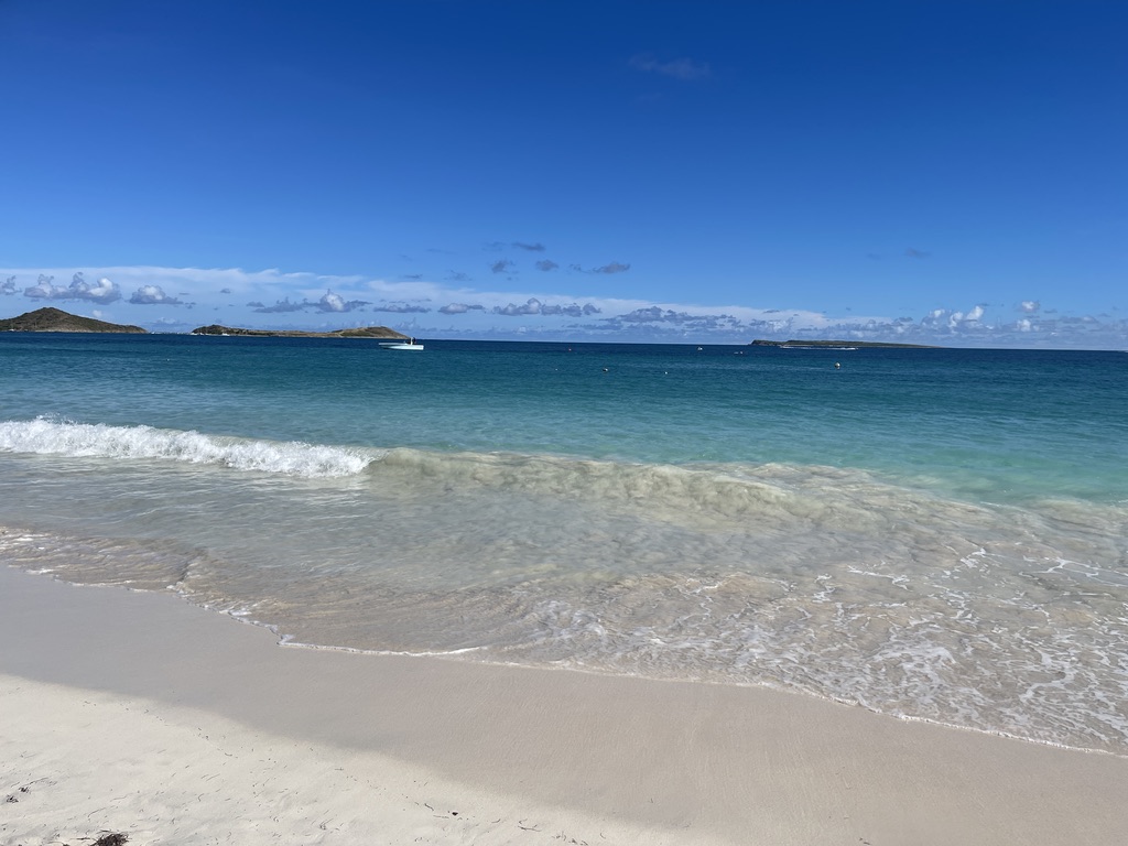 Calm turquoise water washing onto pale sand at Orient Bay, St. Martin, with small islands and a boat on the horizon under a deep blue sky.